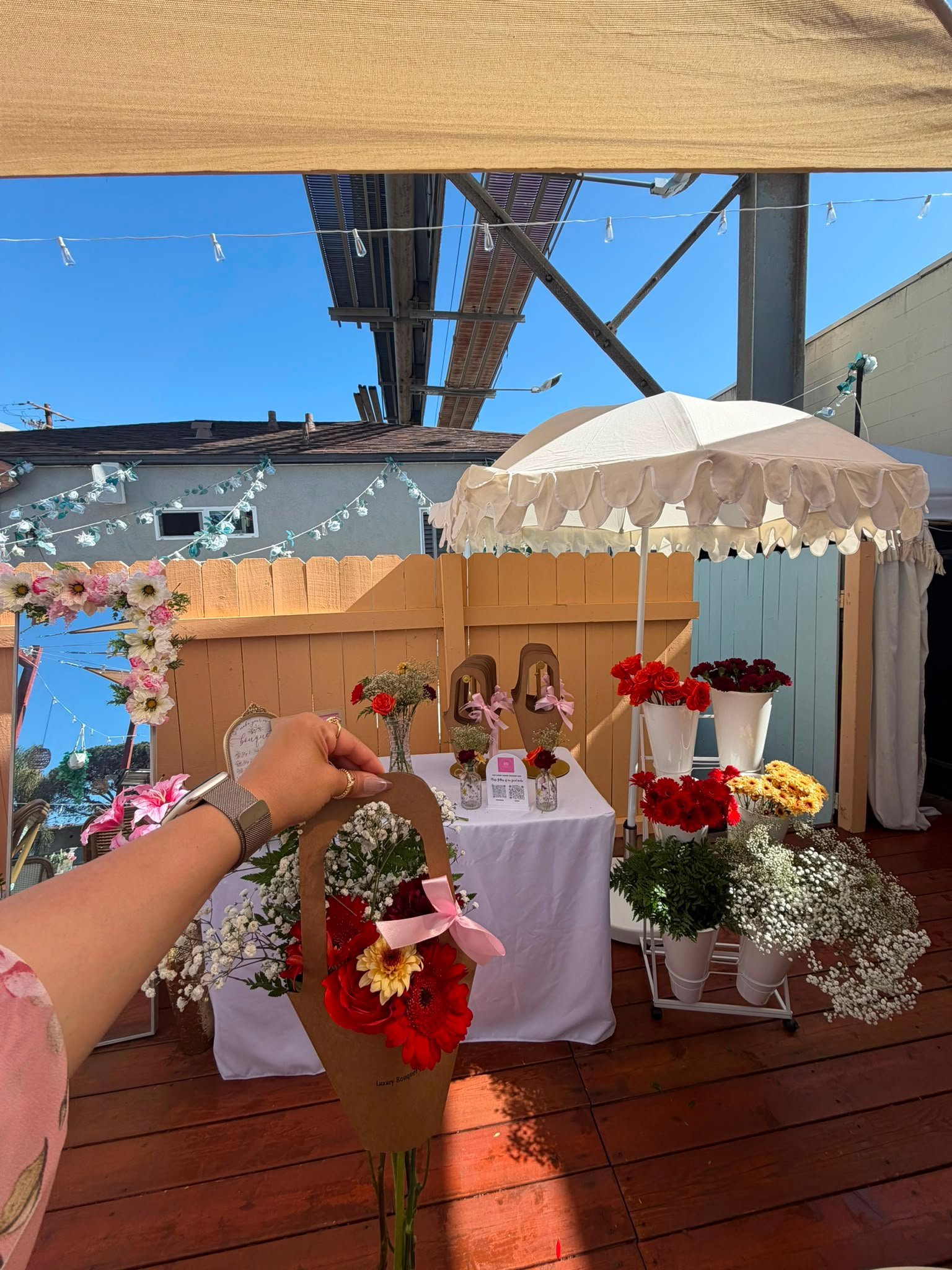 Outdoor rooftop wedding or event setup with colorful flower arrangements, white tent canopy, and decorated wooden fence under blue sky