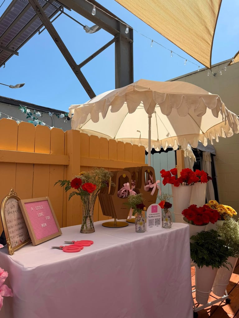 Outdoor event display with pink tablecloth under beige umbrella, decorated with red and pink flowers in vases and signage