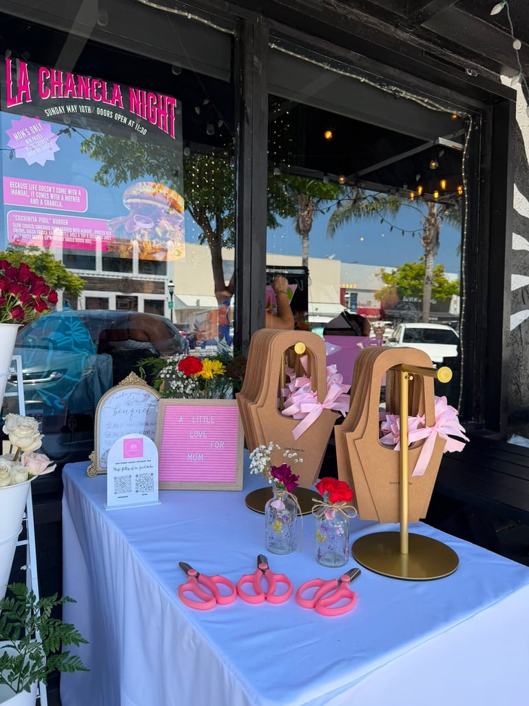 Colorful boutique storefront display with neon pink signage, wooden jewelry stands, and decorative flowers on a blue table