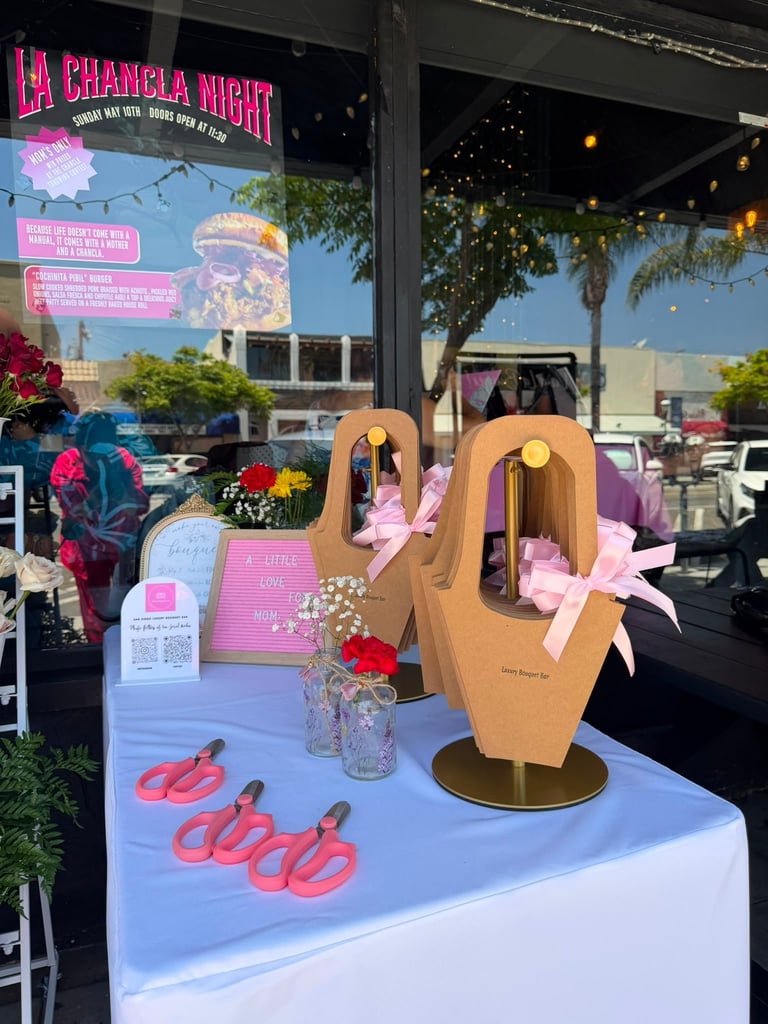 Colorful outdoor event display featuring wooden hand sculptures, pink flamingo floats, flowers, and neon signage for La Chancla Night