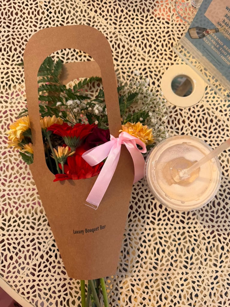 Brown kraft paper flower bouquet with red, yellow and pink flowers tied with pink ribbon, displayed on patterned surface with cosmetic products