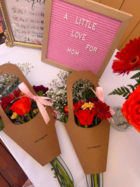 Pink letter board displaying A LITTLE LOVE FOR MOM with red flower bouquets in kraft paper wrapping and white babys breath flowers nearby