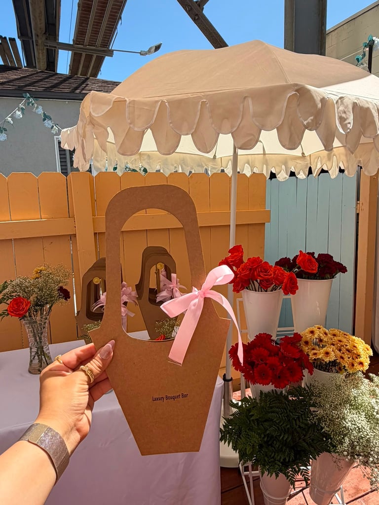 Hand holding a brown decorative flower box with pink bow in front of a flower stall with red and yellow flowers under a beige canopy