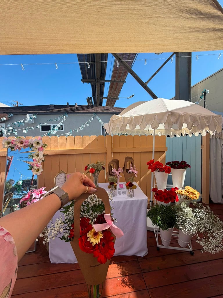 Outdoor rooftop event setup with decorated flower arrangements in pots, white umbrella shade, wooden fence, and beige sail canopy under blue sky