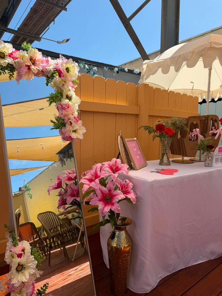 Outdoor event venue with pink and white flower decorations, wooden shade structure, pink tablecloth, and dark wooden chairs under clear blue sky